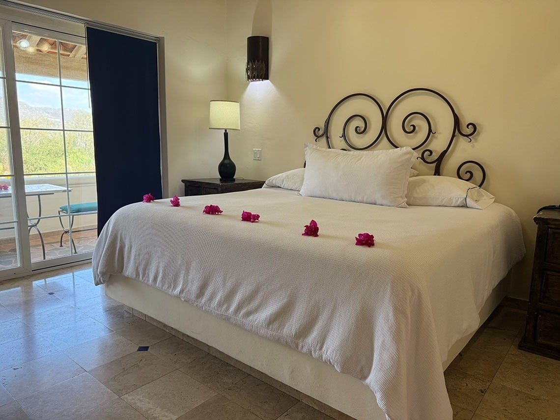 Una habitación luminosa presenta una cama grande con ropa de cama blanca y flores rosas, un cabecero de hierro forjado y una puerta corrediza que da a un balcón con vistas al exterior.