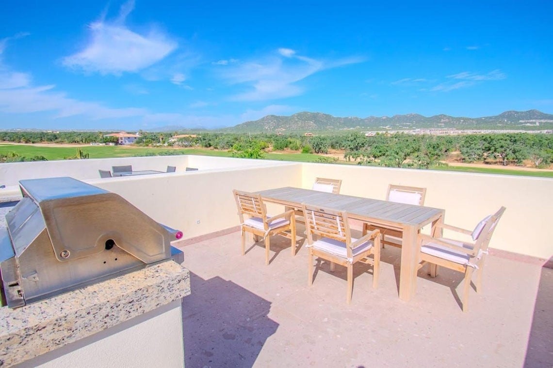 Una terraza al aire libre con una parrilla y un juego de comedor con sillas ofrece vistas a un campo de golf, montañas y un cielo azul brillante.