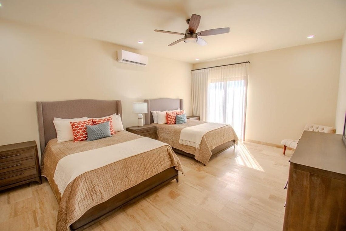 Un dormitorio luminoso con dos camas idénticas, un ventilador de techo, aire acondicionado y una ventana con cortinas transparentes.