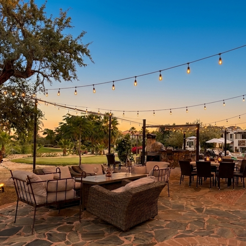 This image captures a spacious outdoor patio area, shaded by light-colored fabric canopies, featuring comfortable lounge seating and dining tables on a distinctive stone and brick mosaic floor, with a golf course visible in the background.