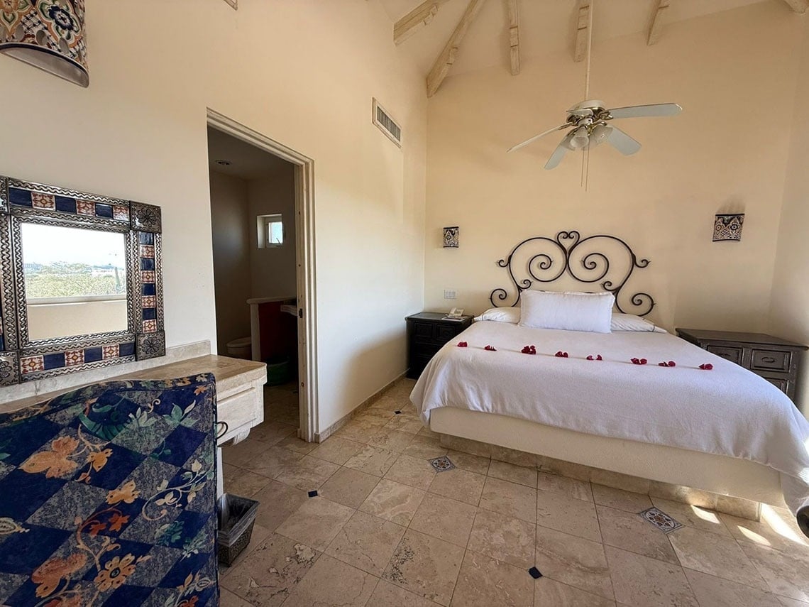 Un dormitorio de estilo rústico y cálido, con una cama adornada con pétalos de rosa, un tocador con espejo y un baño visible a través de una puerta.