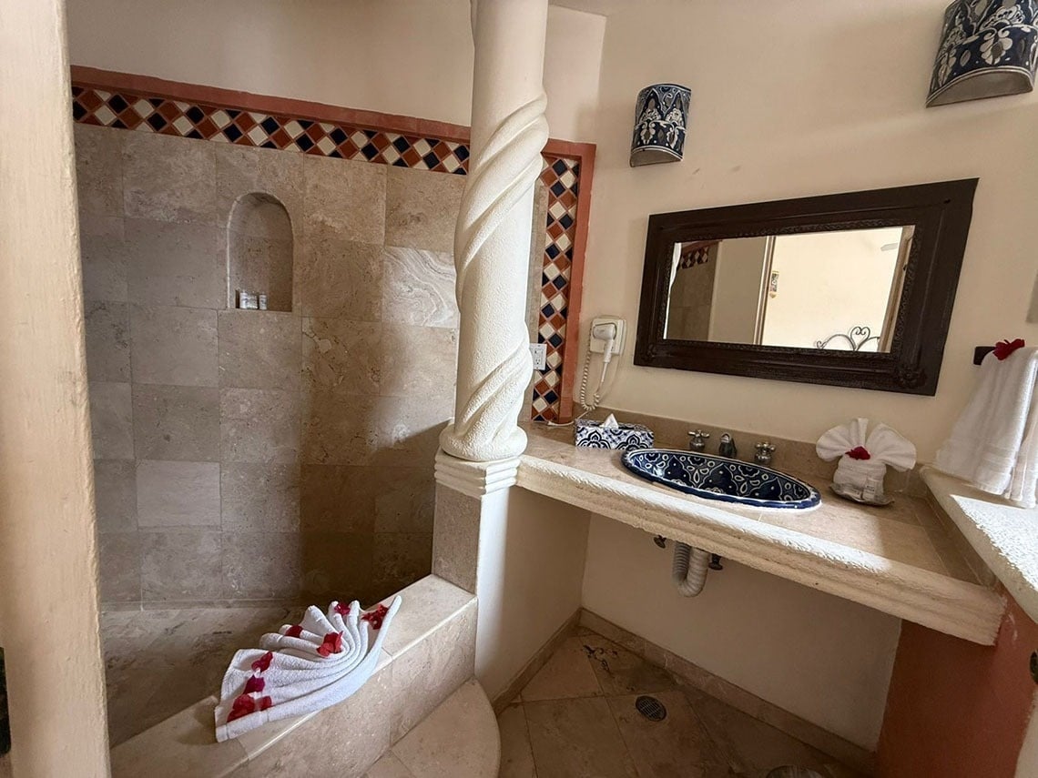 Un baño de estilo tradicional presenta una ducha de azulejos con una hornacina, una columna retorcida, un tocador con lavabo de cerámica azul y toallas artísticamente dobladas.