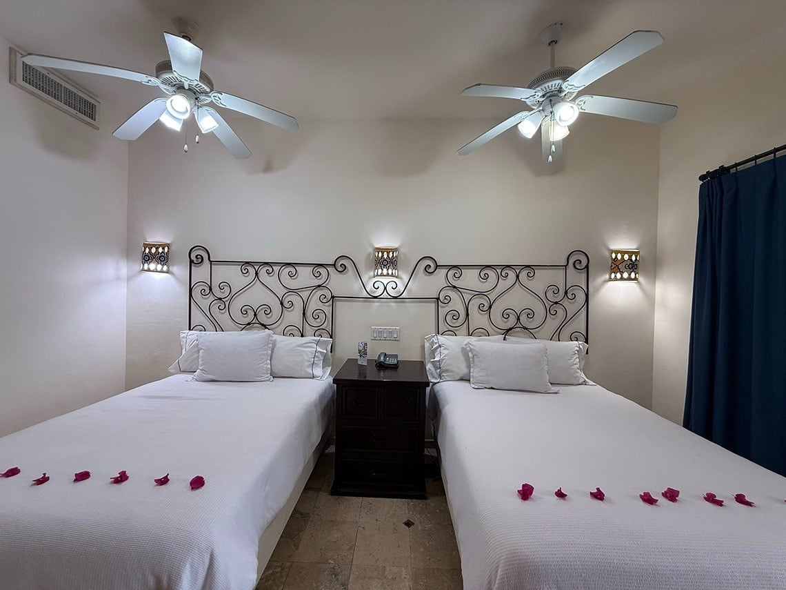 Una luminosa habitación de hotel presenta dos camas individuales con cabeceros de hierro forjado, pétalos de rosa sobre la ropa de cama blanca y ventiladores de techo en lo alto.