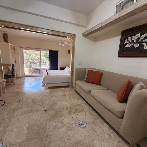 El interior de una estancia con sofá y mesa que conecta con un dormitorio a través de un arco, mostrando una cama y una salida a un jardín.