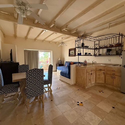 Un espacio diáfano de estilo rústico presenta un comedor con sillas estampadas, una cocina integrada y una sala de estar con sofá azul, todo bajo un techo de vigas de madera y con suelo de baldosas.