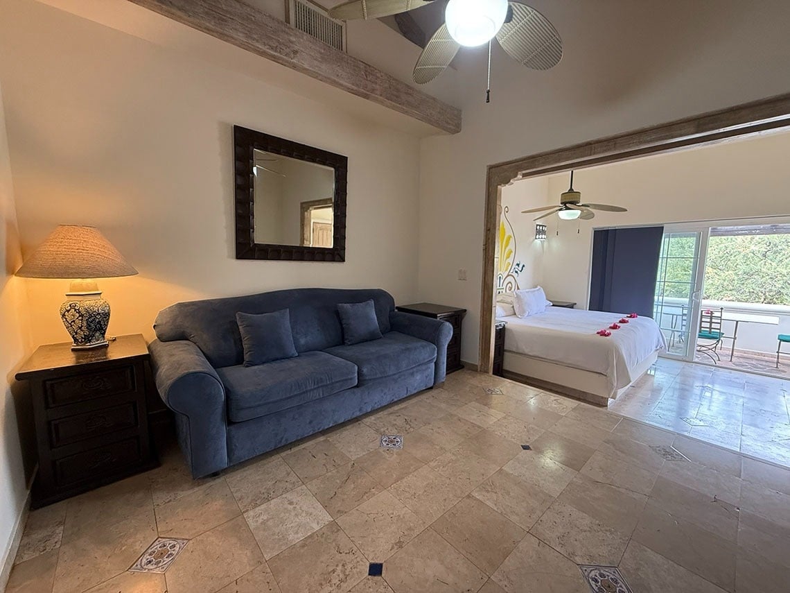 Una luminosa suite de hotel con una sala de estar amueblada con un sofá azul y un dormitorio abierto que cuenta con una cama decorada y acceso a un balcón.