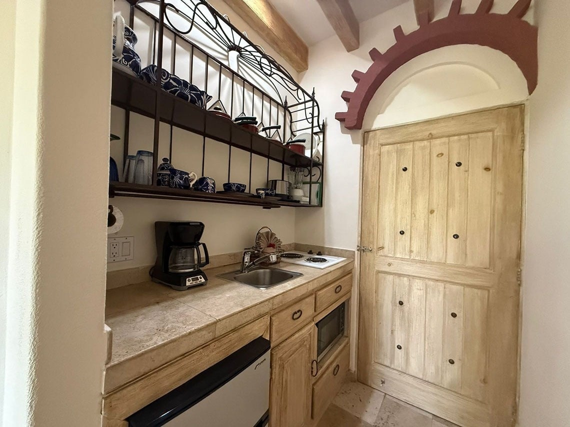 Una pequeña cocina de estilo rústico cuenta con una cafetera, fregadero y microondas bajo estantes de forja llenos de vajilla azul y blanca, junto a una puerta de madera clara enmarcada por un arco decorativo.
