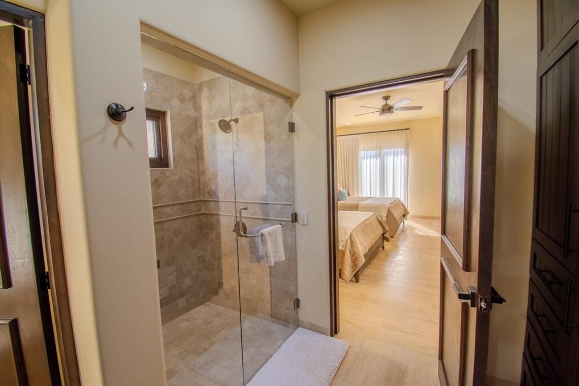 Una vista de un pasillo que conecta una ducha moderna con puertas de cristal y un dormitorio con dos camas, un ventilador de techo y cortinas blancas.