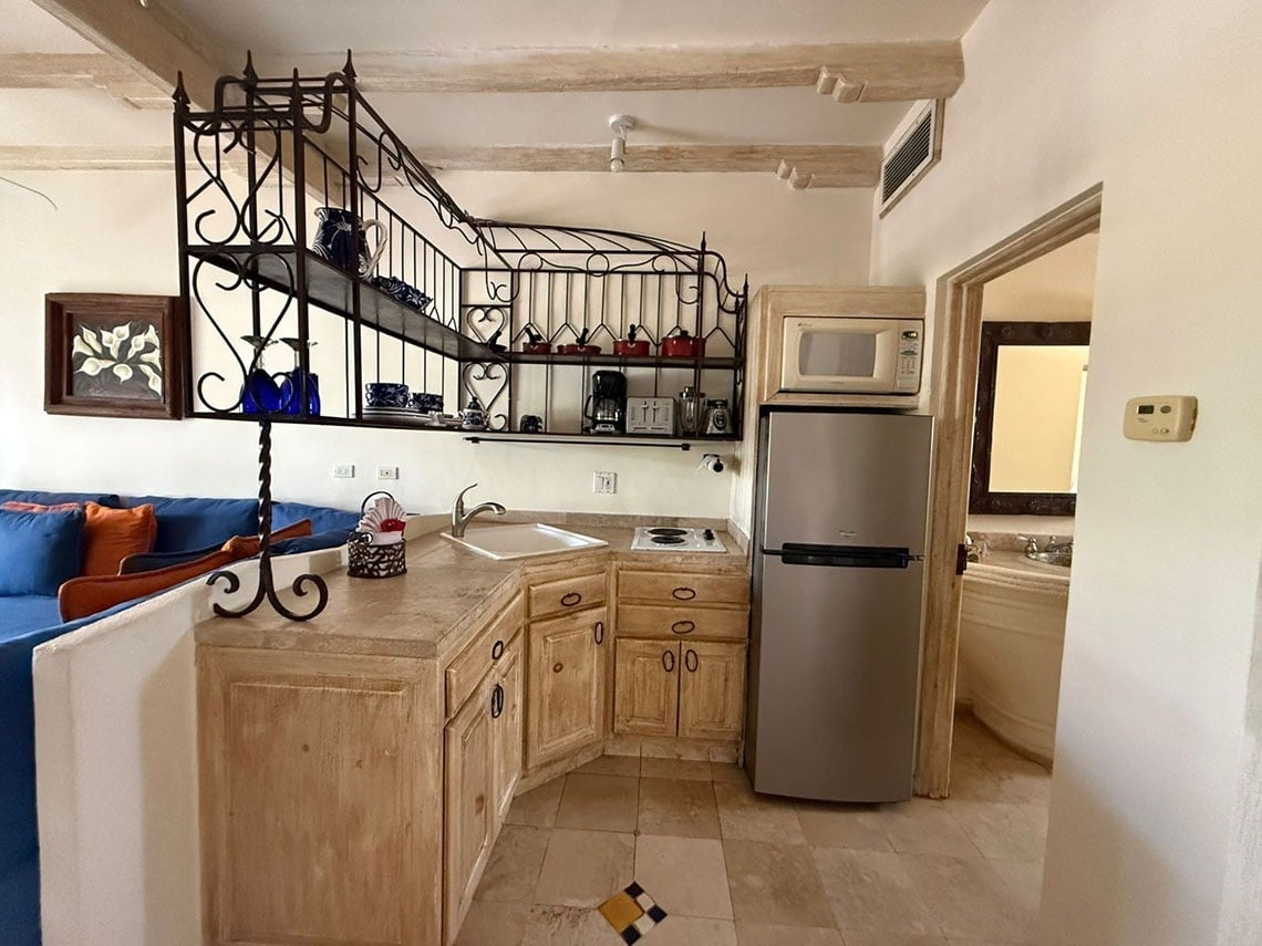 La imagen muestra una cocineta de estilo rústico con encimeras claras, electrodomésticos básicos y estantes ornamentados de hierro forjado, contigua a una sala de estar y un baño.