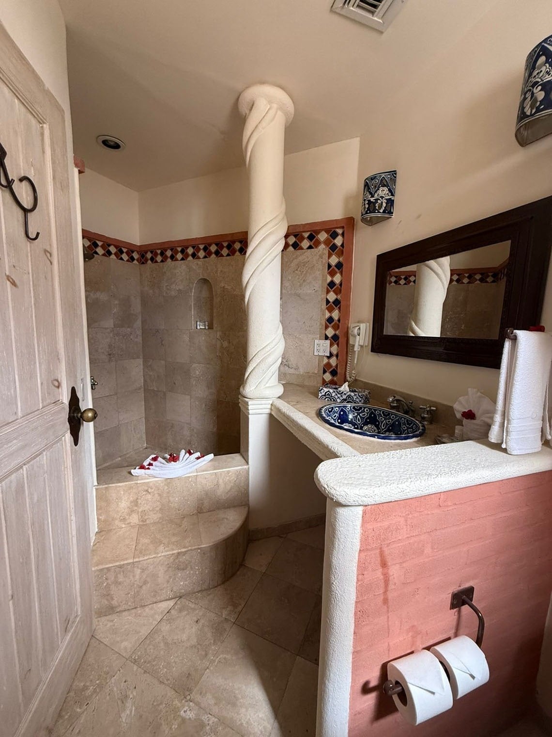 Un baño de estilo rústico presenta una ducha con escalones, un lavabo decorativo azul y blanco, una columna en espiral y una pared de ladrillo pintada de rosa.