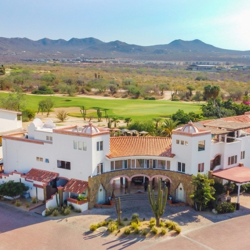 An aerial view captures a bright white resort complex with terracotta roofs, bordered by a green golf course, set against an arid, mountainous landscape under a clear sky.