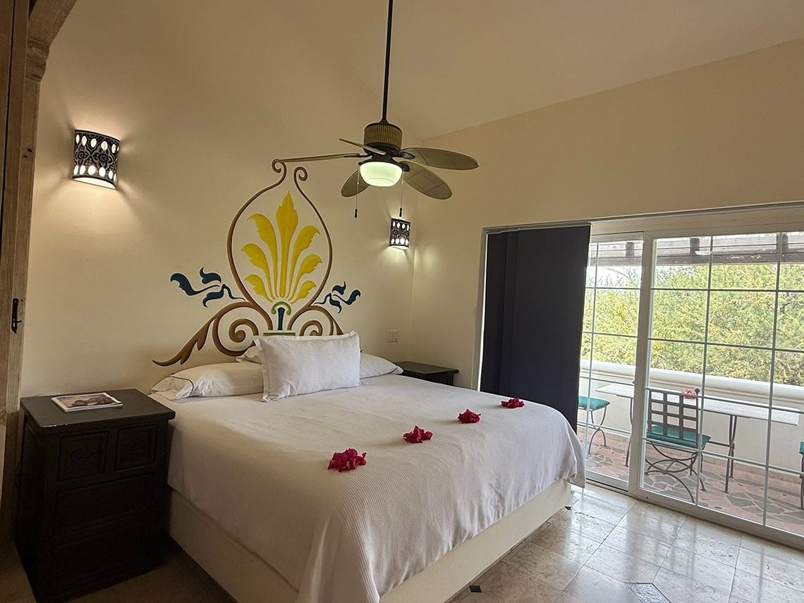 Un dormitorio decorado con un mural floral, una cama con pétalos rojos, un ventilador de techo y un acceso a un balcón con asientos y vistas a la naturaleza.