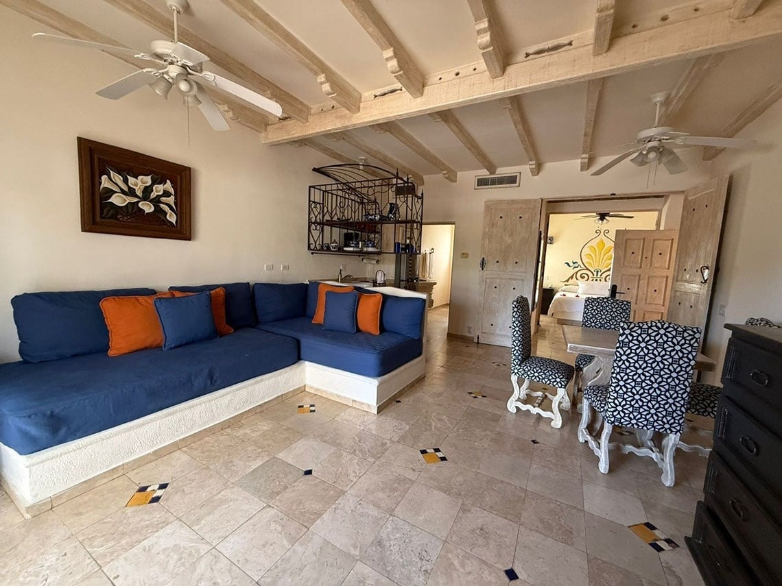 Salón-comedor de estilo rústico con un sofá en L azul, sillas estampadas, techo de vigas de madera y vista a un dormitorio adyacente.