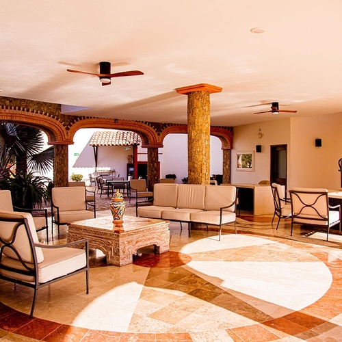 Una espaciosa sala de estar con arcos de piedra, muebles cómodos y vegetación visible a través de aberturas, bañada por la luz del sol.