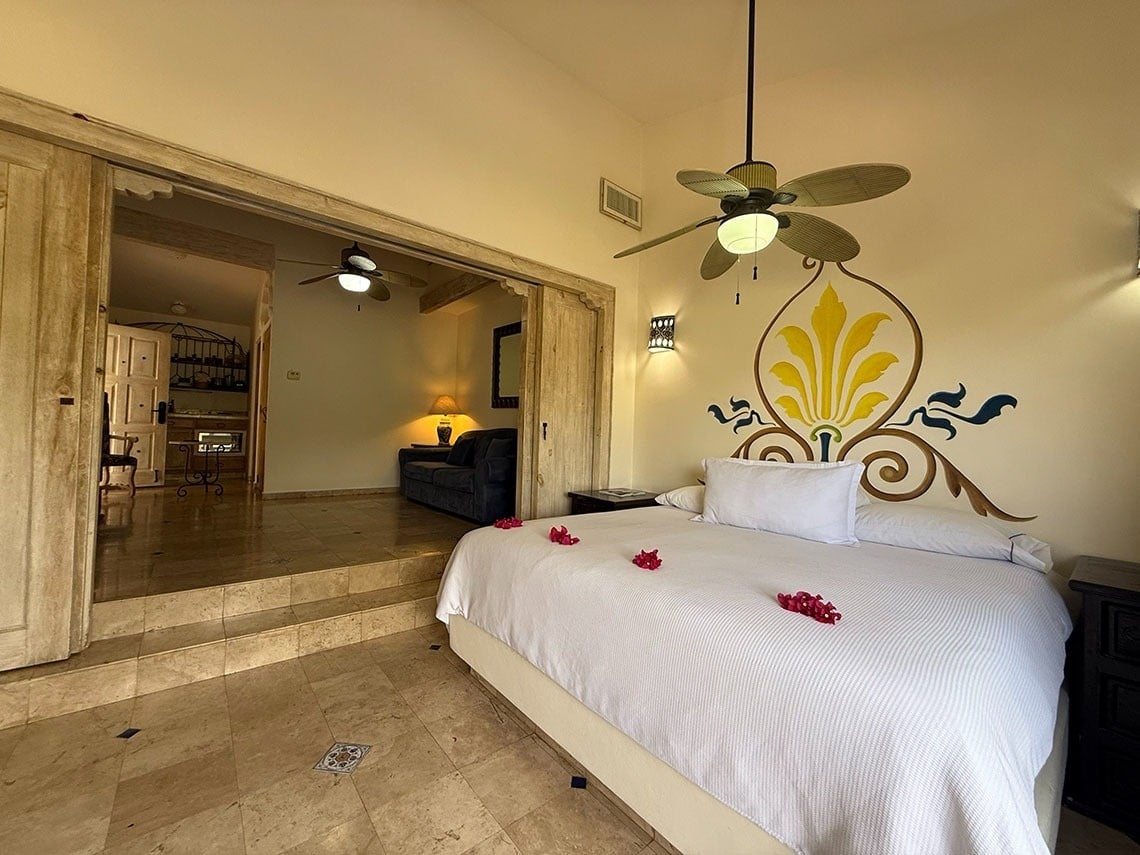 Un dormitorio con una cama decorada con pétalos rojos, un mural y un ventilador de techo, que se abre a una sala contigua elevada por escalones y puertas correderas de madera.