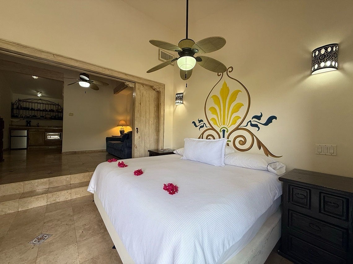 Una acogedora habitación de hotel o suite con una cama grande decorada con flores, un ventilador de techo, arte mural y una cocina/sala de estar adyacente.