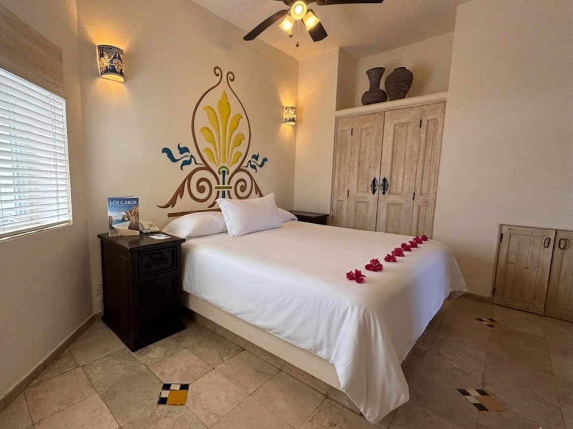 This vibrant bedroom features a bed adorned with rose petals, a colorful wall mural above the headboard, tiled floors, and a window with blinds, creating a warm and inviting atmosphere.