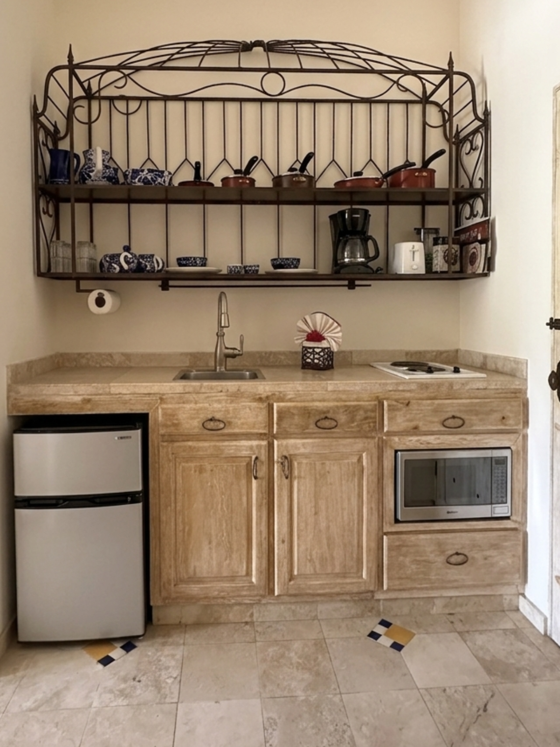 A compact, rustic-style kitchenette features a stovetop, sink, microwave, mini-fridge, and an ornate wrought-iron shelf displaying various kitchen items, all set on beige tiled flooring.