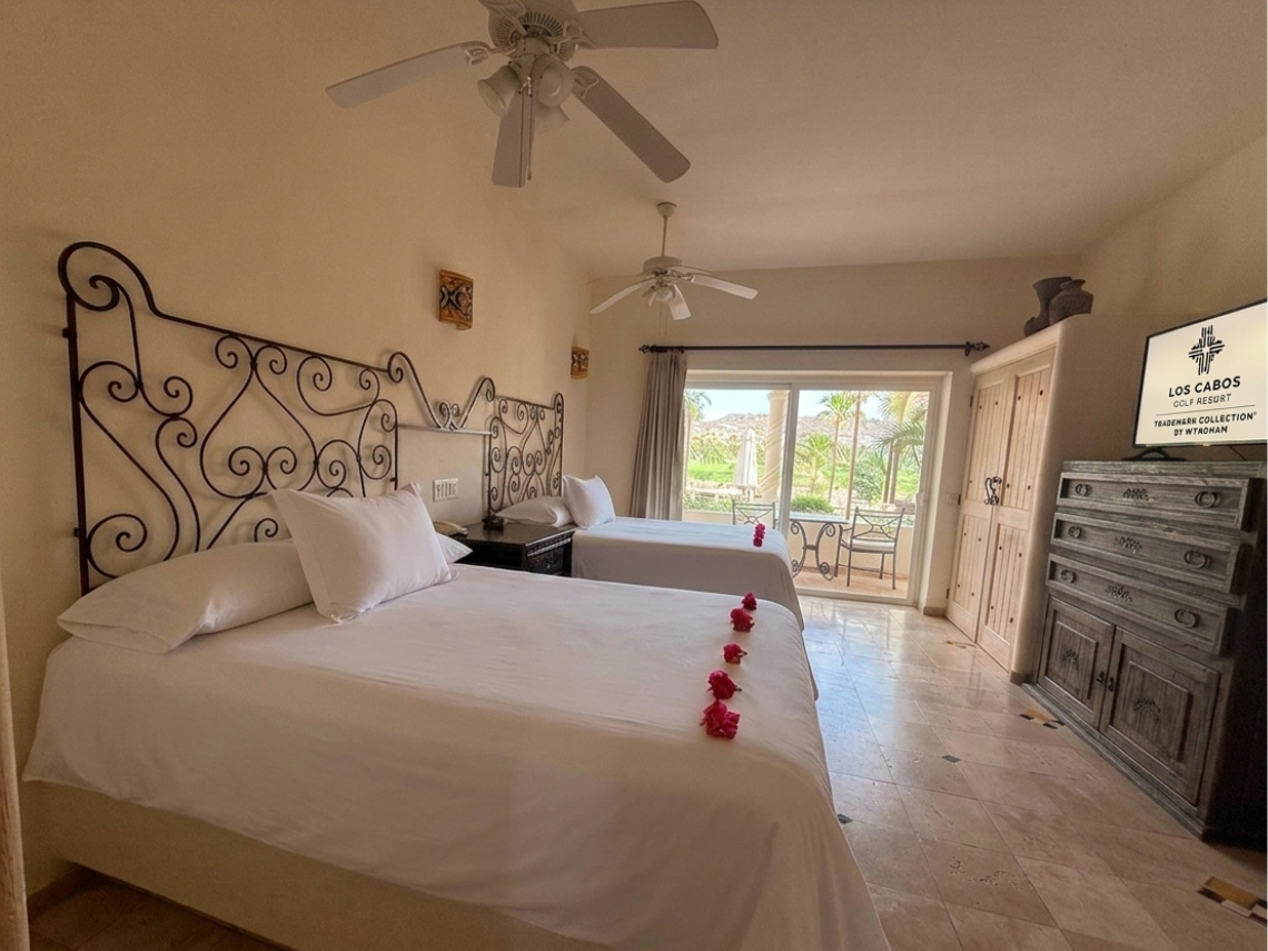 A bright bedroom features two beds with ornate black metal headboards, ceiling fans, a rustic dresser with a television, and a large sliding glass door opening to an outdoor patio.