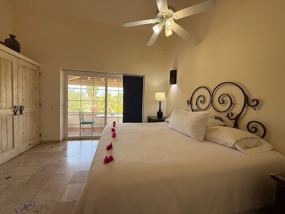 Dormitorio con una cama grande y cabecero de hierro forjado, armario de madera clara, ventilador de techo y puertas corredizas que dan a un balcón con vistas a la vegetación.