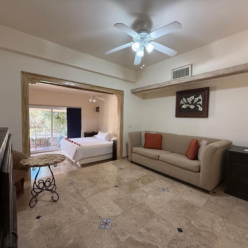 Se observa una sala de estar con sofá y suelo de baldosas que se abre a un dormitorio con cama y pétalos de rosa, con un balcón visible al fondo, a través de un amplio arco.