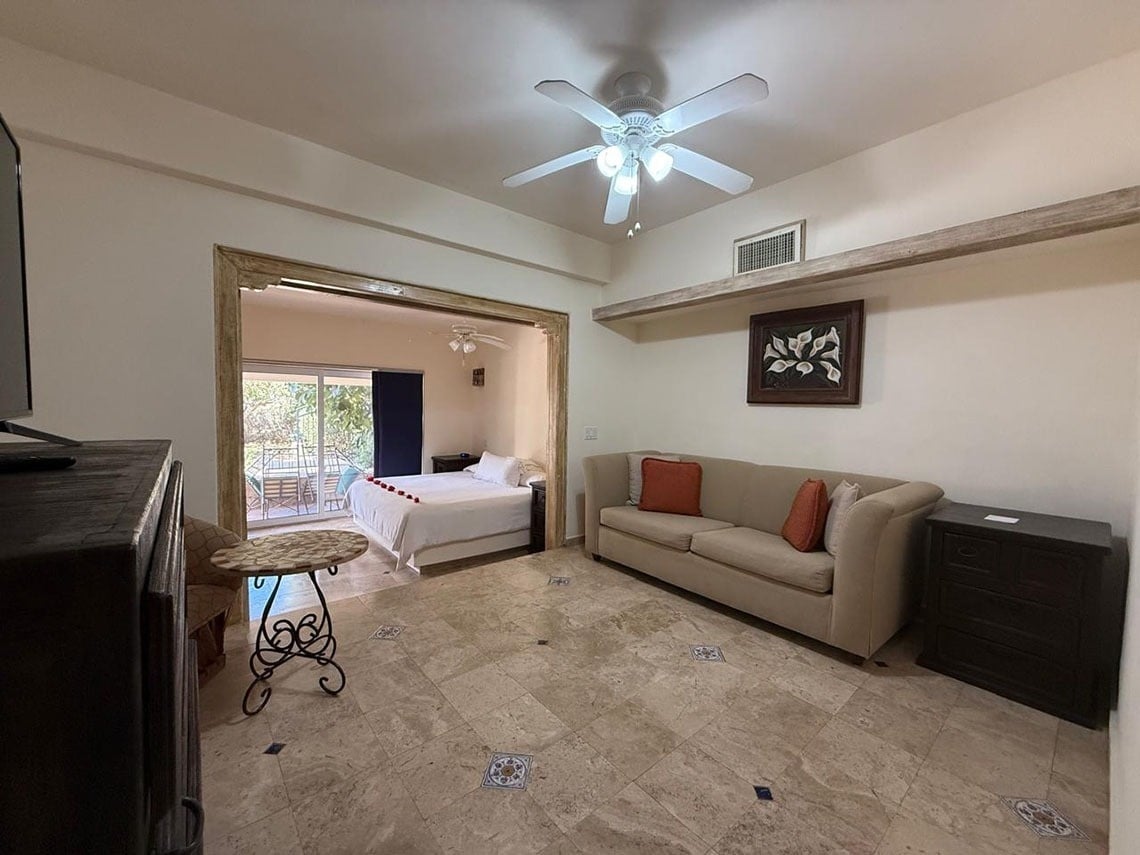 Un espacio abierto combina una sala de estar con sofá y un dormitorio con cama, que da a un balcón, todo con suelo de baldosas.