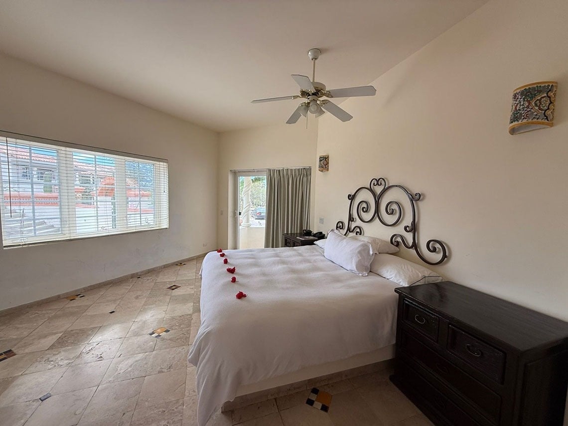 Una luminosa habitación con cama doble, cabecero de hierro forjado, pétalos rojos sobre la cama, ventilador de techo y un gran ventanal con persianas.