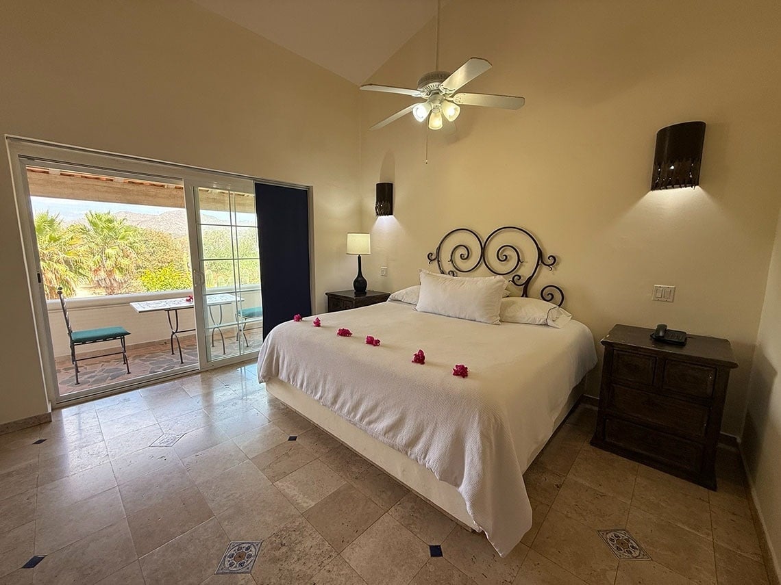 Un dormitorio luminoso con una cama de matrimonio, un balcón con vistas a la vegetación y las montañas, y un suelo de baldosas decoradas.