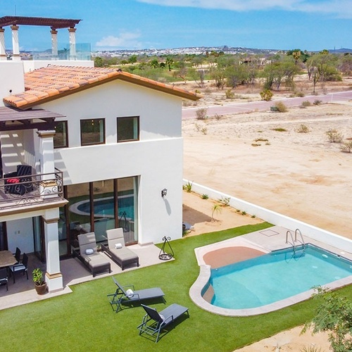 Una casa moderna de color blanco con piscina, césped verde y amplias terrazas, está ubicada en un entorno árido y desértico con vegetación escasa y un camino de tierra.