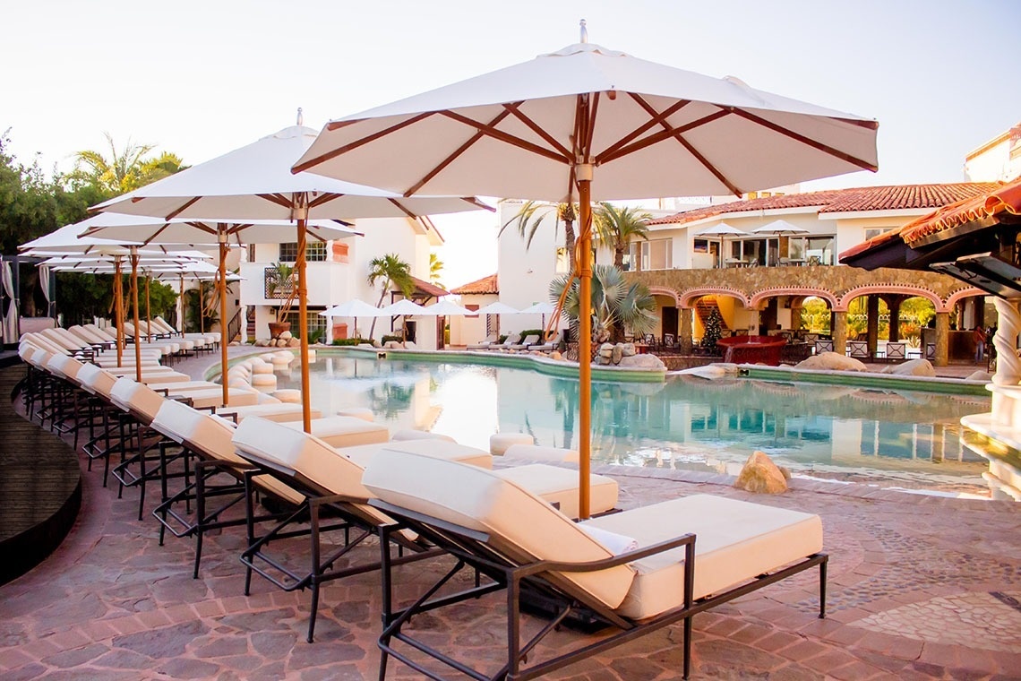 Una soleada piscina de resort rodeada de tumbonas y sombrillas blancas, con arquitectura de estilo mediterráneo al fondo.