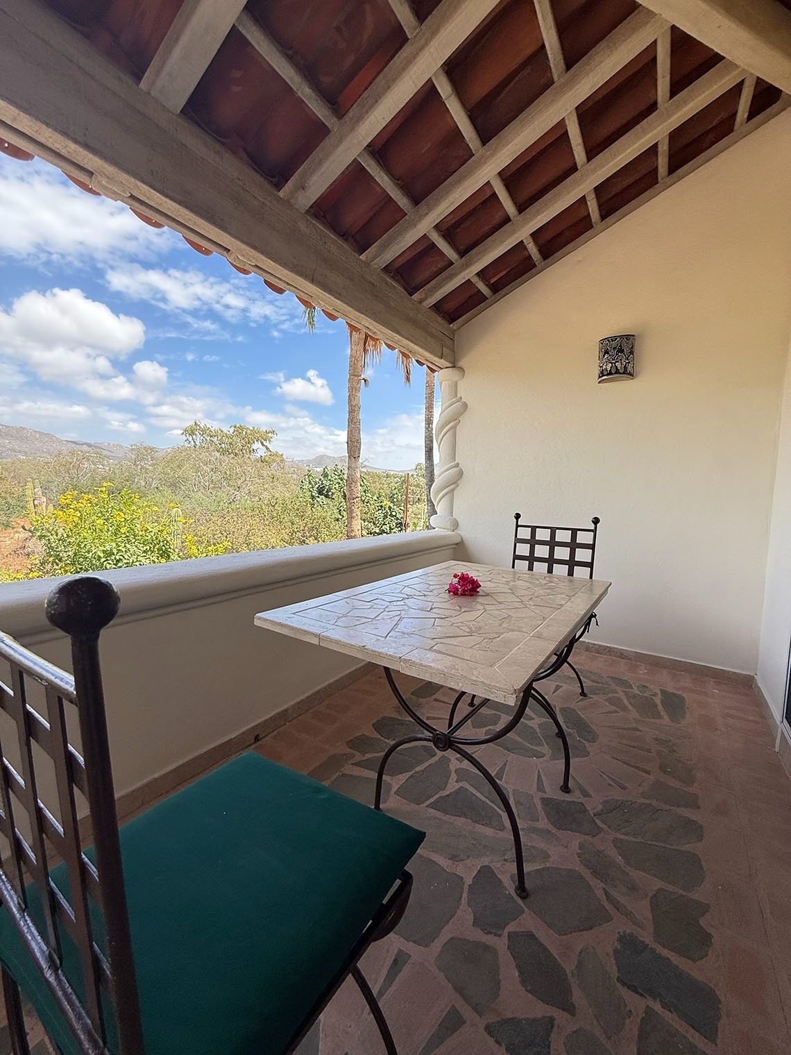 Una terraza rústica con techo de tejas y vigas, amueblada con una mesa y sillas, que ofrece una hermosa vista a un paisaje verde bajo un cielo azul.
