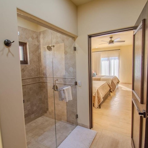 La imagen muestra un pasillo con una ducha de cristal a la izquierda y una puerta abierta a la derecha que revela un dormitorio con dos camas y un ventilador de techo.