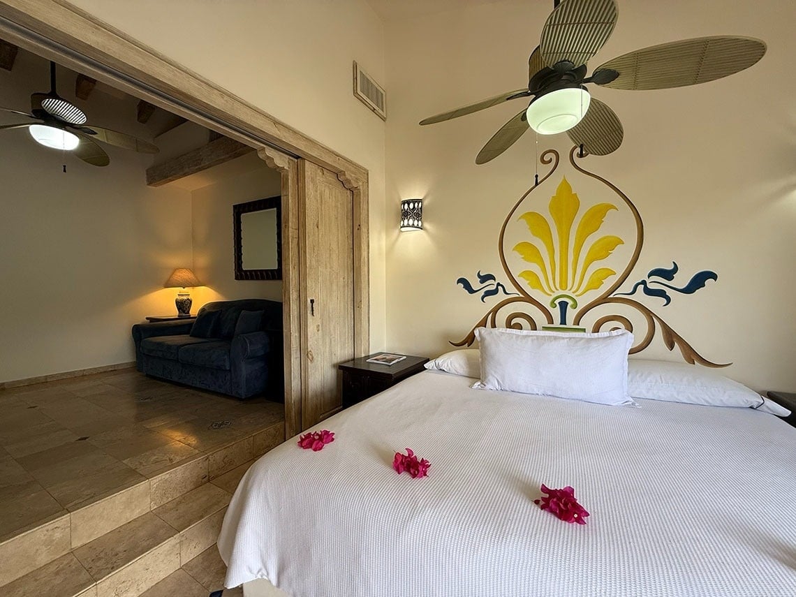 Interior de una habitación dividida en dos niveles, con un dormitorio en primer plano con una cama decorada con flores y un mural, y un área de estar con un sofá en el nivel inferior.