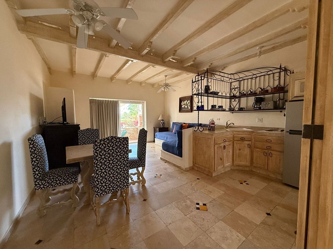 Un estudio o sala de estar de estilo rústico con techos de vigas, que incluye un área de comedor con sillas estampadas, una cocina integrada con gabinetes de madera clara y estantes de hierro forjado, y un suelo de baldosas, con una puerta corredera que da a un balcón.