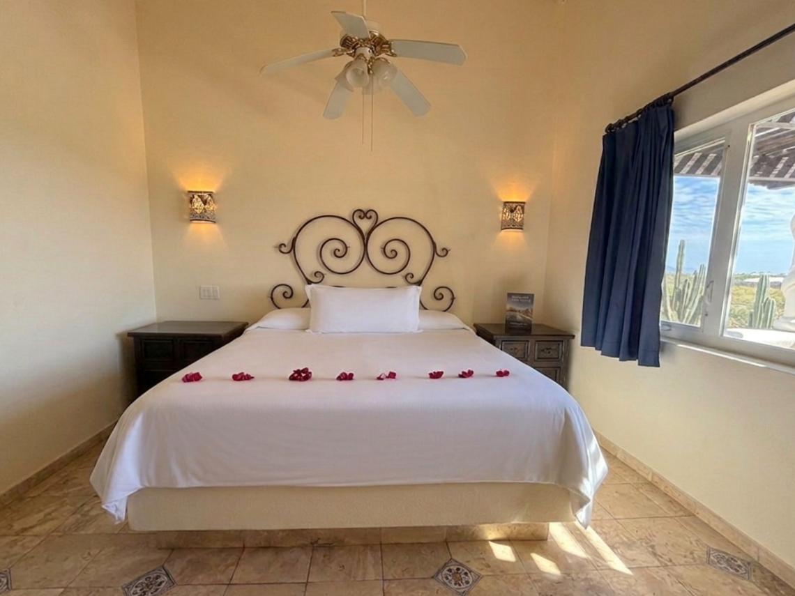 A sun-drenched bedroom features a large bed with a wrought iron headboard and scattered rose petals, offering a wide window view of a sunny outdoor landscape.