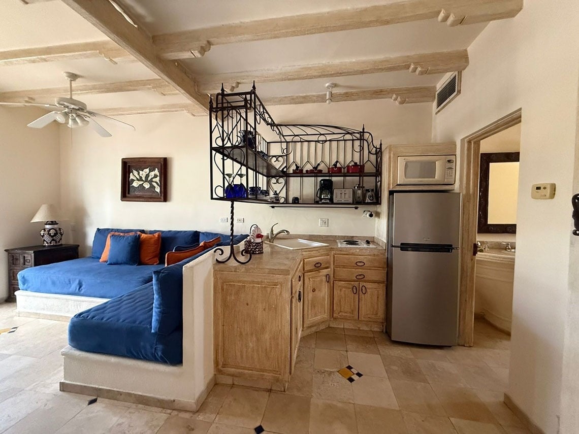 Una sala de estar y cocina de estilo rústico con un sofá azul en forma de L, una estantería de hierro forjado y vigas de madera en el techo.