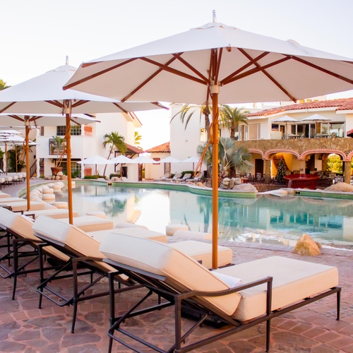 An aerial view showcases a Mediterranean-style resort complex with red-tiled roofs and a large, winding swimming pool, situated in an arid coastal landscape with distant mountains and a city.