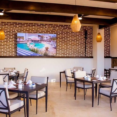 Un comedor de restaurante de estilo rústico-moderno, con mesas y sillas de metal y cojines claros, destaca por una pared de ladrillo visto con una gran televisión y lámparas colgantes de mimbre.