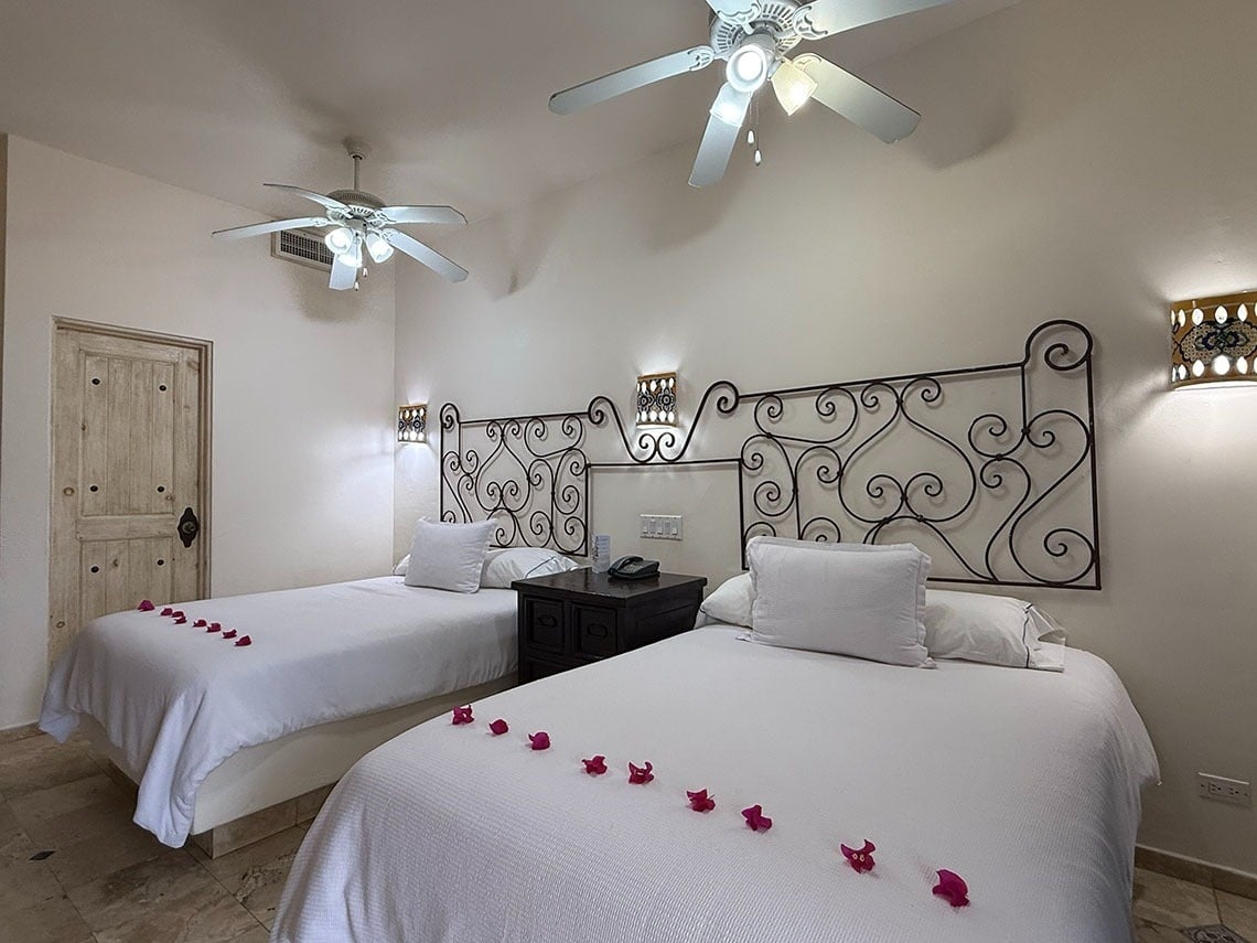 Una habitación de hotel presenta dos camas con ropa de cama blanca y pétalos de flores, cabeceros de hierro forjado y ventiladores de techo.