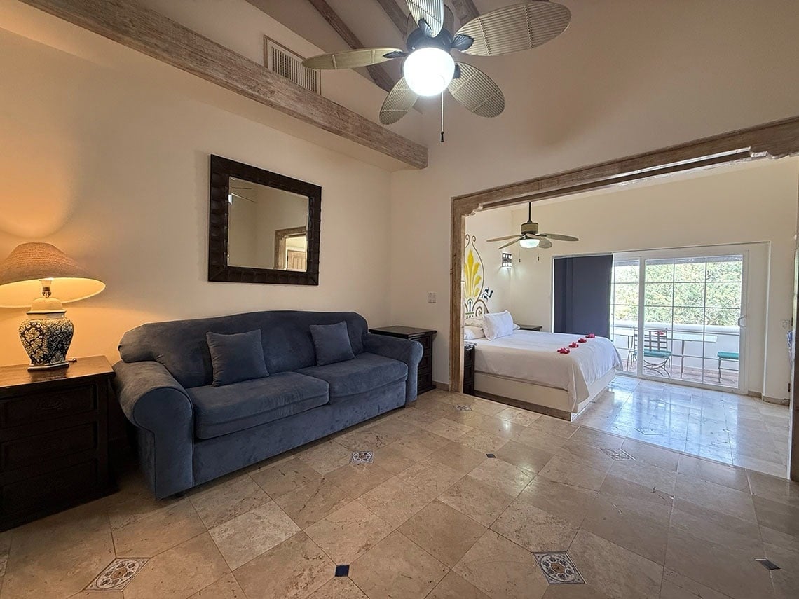 Un espacio interior dividido en una sala con sofá y un dormitorio con cama y balcón, todo sobre un suelo de baldosas.