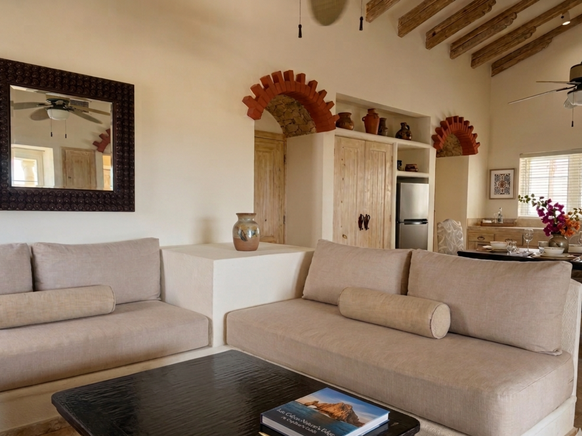 This spacious open-concept living area features a blue sofa and an orange armchair, a dining table with patterned chairs, and a visible kitchen in the background, all set on light tiled floors under a beamed ceiling with ceiling fans.