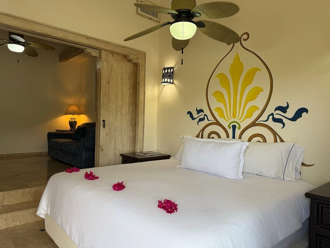 Un dormitorio con una cama grande adornada con pétalos rosados, un mural decorativo en la pared, un ventilador de techo y una sala de estar adyacente visible a través de una puerta de madera.