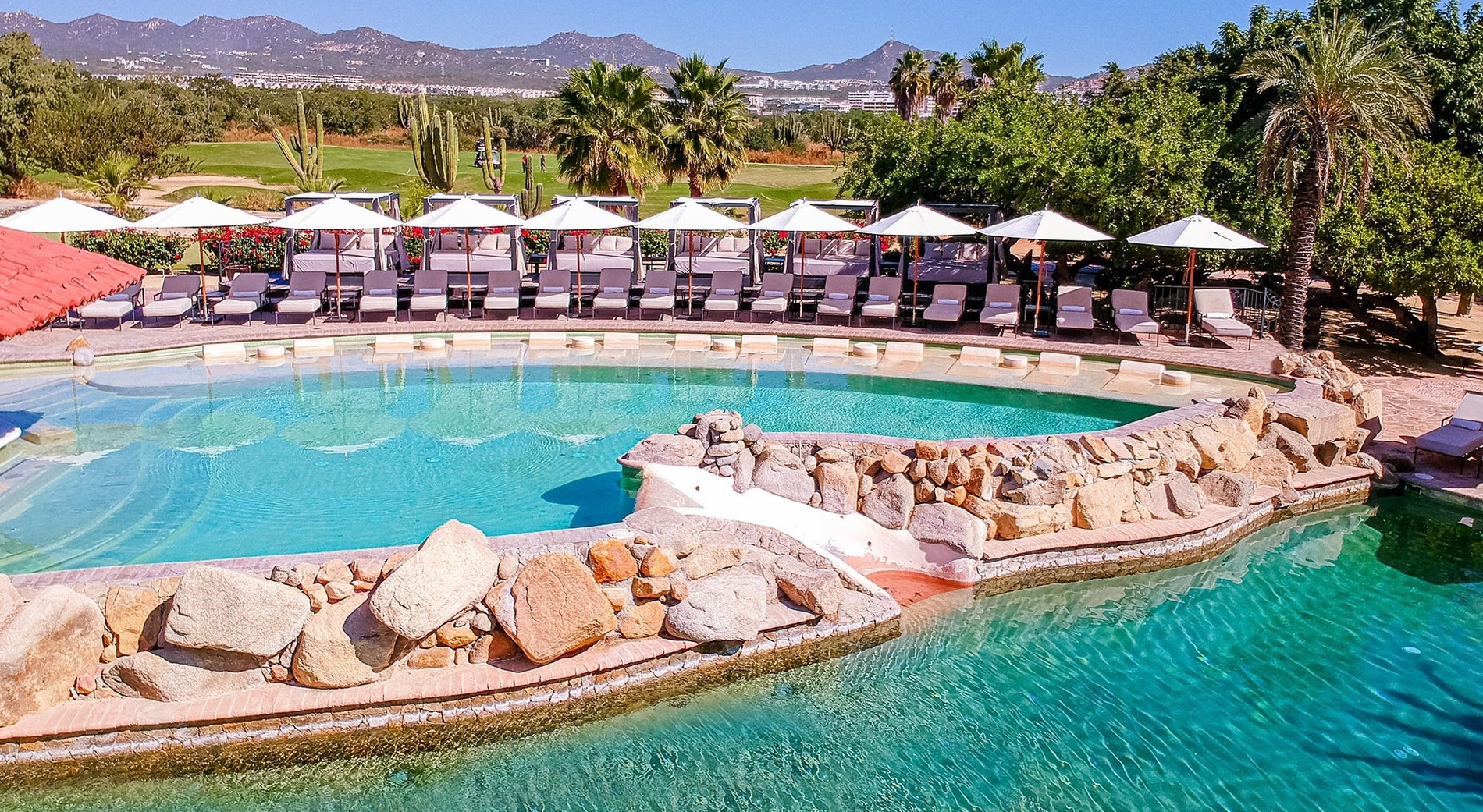 Una piscina de resort con tumbonas y sombrillas, elementos rocosos y un campo de golf con montañas y vegetación desértica al fondo.