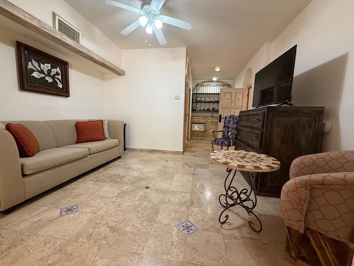 Una acogedora sala de estar con sofá beige, TV, muebles de madera rústicos y piso de baldosas con detalles decorativos, se extiende hacia una pequeña área de cocina al fondo.