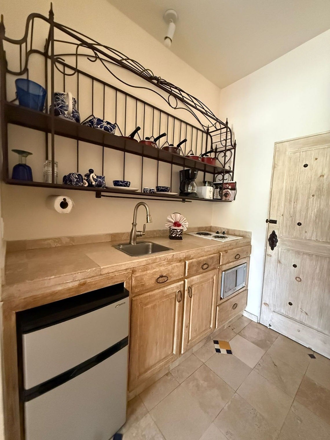 Una cocineta rústica con gabinetes de madera clara, encimera de piedra, electrodomésticos compactos y estanterías de hierro forjado repletas de vajilla y utensilios de cocina.