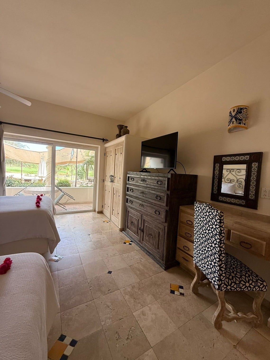 Un dormitorio luminoso con dos camas, un mueble de madera oscura con televisor, un tocador claro con espejo y una silla estampada, y un ventanal que da a un patio exterior, todo con suelo de baldosas claras y detalles coloridos.