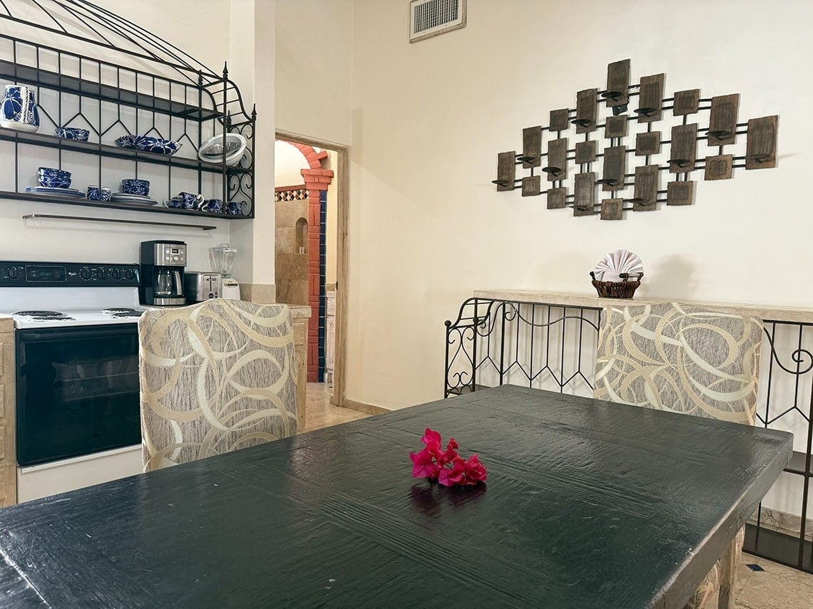 Este comedor y cocina presenta una mesa oscura con flores rosadas, sillas tapizadas, una estantería de hierro forjado con vajilla azul y una gran decoración de pared de madera y metal.