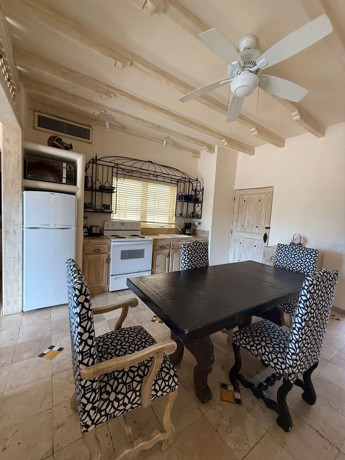 Una cocina-comedor de estilo rústico presenta electrodomésticos blancos, muebles de madera clara, una mesa oscura con sillas tapizadas en blanco y negro, y un techo con vigas expuestas.