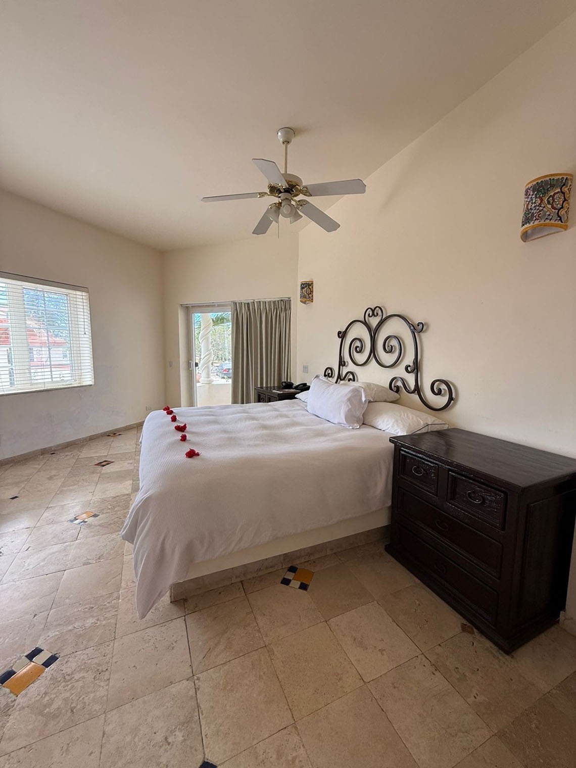 Un dormitorio luminoso con una cama de sábanas blancas y un cabecero de hierro forjado, suelo de baldosas claras con detalles de colores, un ventilador de techo y una puerta con cortinas que da al exterior.