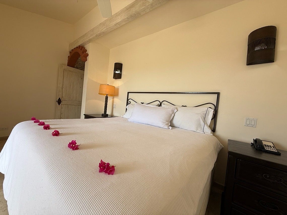 Un dormitorio cálido con una cama blanca adornada con flores rosas, mesitas de noche con lámparas y una puerta rústica bajo un arco.